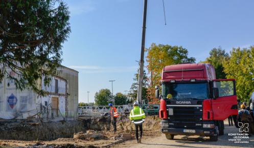 Plac budowy. Po lewej stronie jest wykopany dół, pod budowę fundamentów. Obok niego stoi prostokątny budynek. Po prawej stronie stoi czerwony tir. Za nim widać ramię żurawia budowlanego. 