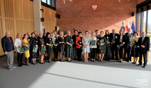 Sala konferencyjna. Dwadzieścia sześć osób stoi obok siebie w rzędzie – sześciu mężczyzn i dwadzieścia kobiet. Większość w rękach trzyma dyplom w zielonej teczce i białą różę.