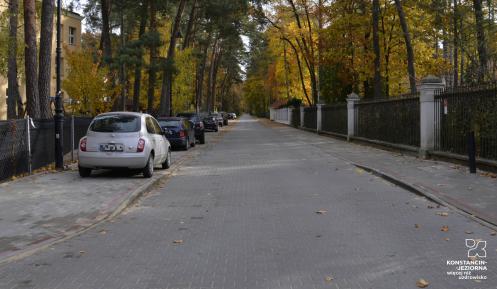 Ulica z kostki brukowej. Po jej lewej stronie stoją zaparkowane w rzędzie samochody. Po prawej stronie znajduje się chodnik. W dali widać wysokie drzewa z żółtymi i brązowymi liśćmi.