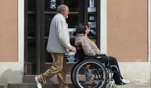  Miasto. Po chodniku idzie mężczyzna, który pcha wózek inwalidzki. Na wózku siedzi kobieta. W tle drzwi budynku. 