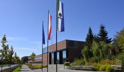 Budynek Urzędu Miasta i Gminy Konstancin-Jeziorna wykonany z czerwonej cegły, przed budynkiem stoją trzy maszty z flagami: Polski, Unii Europejskiej i Konstancina-Jeziorny. Po prawej stronie drzewa i krzewy, po lewej – droga z samochodami. 