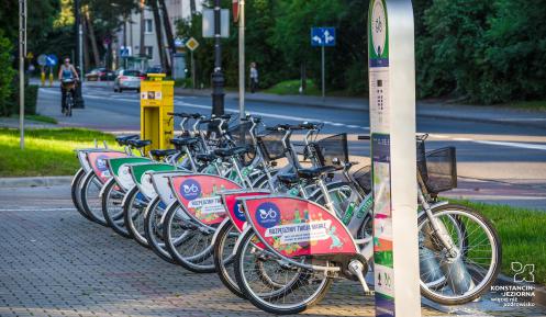 Stacja wypożyczalni rowerów miejskich z kilkunastoma rowerami. W tle ulica oraz jadąca po chodniku rowerzystka.