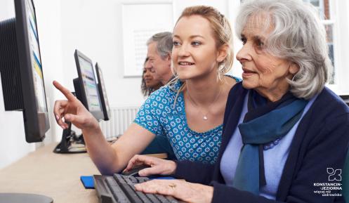 Sala komputerowa. Młoda kobieta pokazuje palcem coś na ekranie komputera starszej osobie (kobiecie), która trzyma ręce na klawiaturze komputerowej.