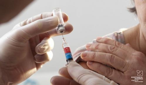 Zdjęcie. Po lewej stronie widoczne są dłonie z białych rękawiczkach. Po prawej stronie, starsza Pani odsłania ramie do wykonania zastrzyku. 