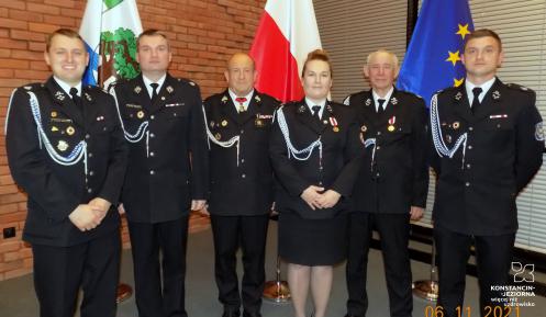 Sześć osób (pięciu mężczyzn i kobieta) w mundurach strażackich stoją w rzędzie na tle flag Polski, Konstancina-Jeziorny i Unii Europejskiej.