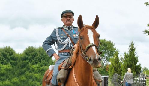 Mężczyzna ubrany w strój wojskowy siedzi na brązowym koniu. W tle zielone drzewa.