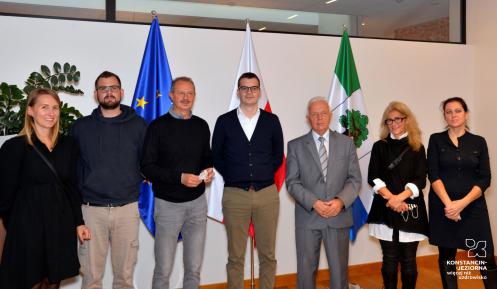 Sala konferencyjna. Siedem osób stoi obok siebie, trzy kobiety i czterech mężczyzn. W tle biała ściana oraz trzy flagi: flaga Konstancina-Jeziorny, flaga Polski oraz flaga Unii Europejskiej.