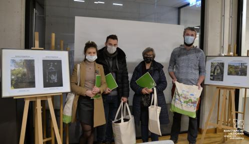 Sala konferencyjna. Cztery osoby w maseczkach stoją przodem i trzymają w rękach torby płócienne oraz zielone teczki na dokumenty.