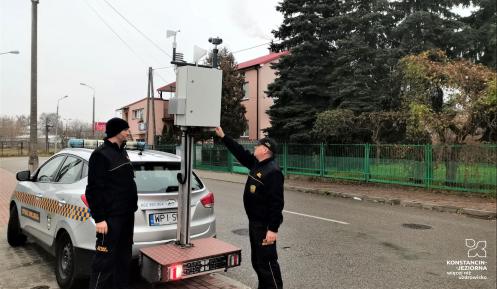 Samochód osobowy, do którego haku przymocowana jest platforma z czujnikiem jakości dymu. Urządzenie obsługują dwaj strażnicy miejscy. Samochód zaparkowany jest na chodniku. W tle widać ulicę i budynek mieszkalny. 