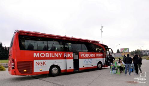 Na chodniku stoi duży czerwony autobus z białym napisem z boku Mobilny Punkt Poboru Krwi. Przed autobusem stoi sześć osób. 