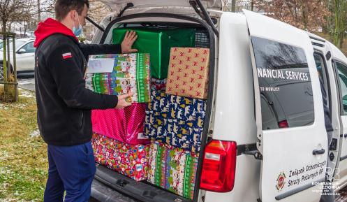 Samochód dostawczy, który ma otwarty bagażnik. Znajdują się w nim duże pudła – prezenty świąteczne. Obok samochodu stoi mężczyzn, który pakuje do niego kolejną paczkę.