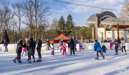 Przestrzeń miejska, ludzie w strojach zimowych jeżdżą na lodowisku, w tle park i dach amfiteatru.