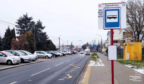 Ulica. Po prawej stronie znajduje się chodnik. Na nim stoi wysoki słup z tabliczką – nazwą przystanku autobusowego i rozkładem jazdy. 