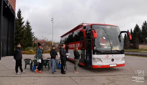 Na chodniku stoi czerwony autokar, z boku jest duży napis Mobilny punkt poboru krwi, obok jest godło Polski i napis Minister Zdrowia. Przed autokarem w kolejce stoi pięć osób.