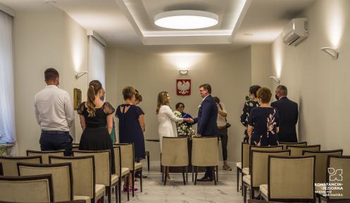 Sala ślubów cywilnych. Na środku naprzeciwko siebie stoi para młoda – trzymają się za ręce, składają przysięgę. Po bokach sali stoją goście – około 10 osób. 