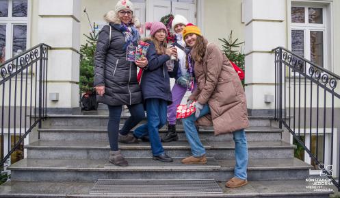 Na schodach przed budynkiem stoją cztery osoby z puszkami WOŚP oraz nalepkami – czerwonymi serduszkami WOŚP.