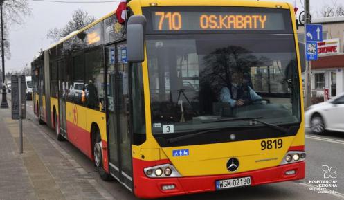 Ulica, po której jedzie czerwono-pomarańczowy autobus komunikacji miejskiej. Nad jego przednią szybą wyświetla się napis: 710 Osiedle Kabaty.