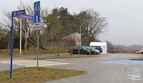 Parking z kostki brukowej. Poi lewej stronie widoczny drogowskaz z nazwami ulic. W tle kilka zaparkowanych samochodów. Na końcu parkingu stoi biały duży namiot. 