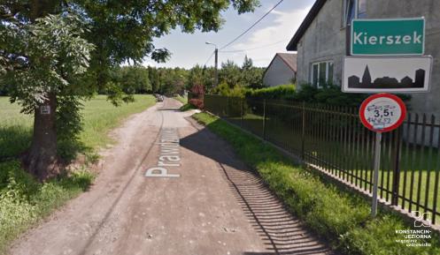 Polna droga, po prawej stronie znak drogowy z nazwą miejscowości Kierszek i zabudowania mieszkalne i gospodarcze, w tle drzewa.
