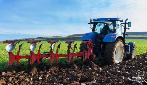 Niebieski traktor, do którego tyłu dołączony jest czerwony pług obrotowy orzący ziemie na polu. W tle widać las. 