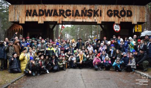 Grupa kilkuset osób w strojach harcerskich na tle drewnianej bramy wjazdowej z napisem: Nadwarciański Gród.