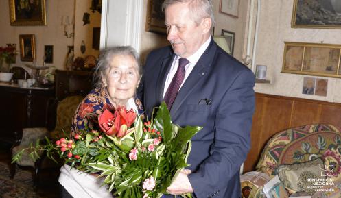 Stojący po prawej stronie mężczyzna w średnim wieku, ubrany w garnitur, wręcza bukiet kwiatów starszej kobiecie stojącej po lewej stronie. Kobieta patrzy na wprost. Za nimi widać mieszkanie, ze ścianami udekorowanymi licznymi obrazami.