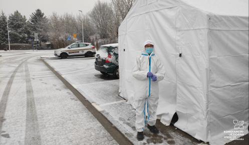 Parking samochodowy z kostki brukowej. Po prawej stronie widoczny biały duży namiot, przed którym stoi osoba w białym kombinezonie i maseczce, z tyłu stoją dwa samochody osobowe.