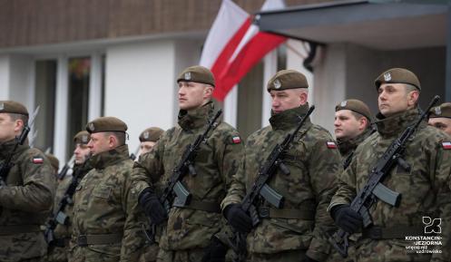 Grupa kilkunastu żołnierzy w umundurowaniu i z bronią automatyczną przewieszoną na piersiach, na głowach berety, za nimi widoczny fragment budynku z umocowanymi dwiema flagami biało-czerwonymi.