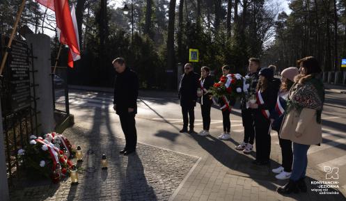 Zdjęcie przedstawia sytuację opisaną w artykule. Na pierwszym planie stoi mężczyzna z głową pochyloną w stronę lewą, gdzie widoczny jest fragment tablicy pamiątkowej, przed tablicą leżą wiązanki kwiatów w kolorach biało-czerwonych oraz znicze, po prawej stronie w rzędzie stoi piątka młodzieży z szarfami biało czerwonymi przepasanymi przez piersi, jeden chłopak trzyma wiązankę kwiatów, po lewej stronie rzędu stoi drugi mężczyzna, po prawej stronie stoi kobieta.