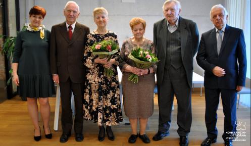 Sala konferencyjna. Trzy pary seniorów stoją obok siebie w rzędzie, kobiety w rękach trzymają bukiety białych kwiatów. 