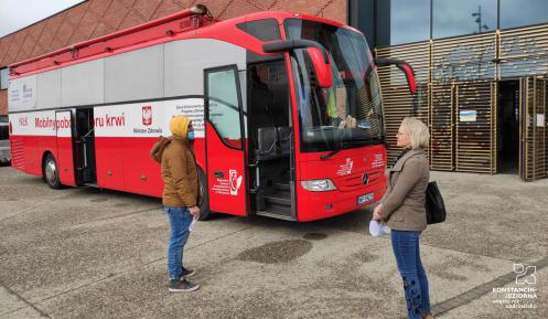  Duży czerwony ambulans stoi przed budynkiem z czerwonej cegły. Jego drzwi są otwarte. Przed wejściem do niego stoją kobieta i mężczyzna. 