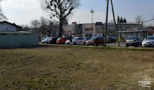 Przestrzeń miejska. Na pierwszym planie fragment działki, w tle parking samochodowy.