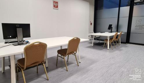 Pomieszczenie z dwoma stołami, na których stoi po komputerze, a przed każdym stołem po dwa krzesła. 