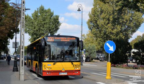 Ulica, po której jedzie czerwono-pomarańczowy autobus komunikacji miejskiej. Nad jego przednią szybą wyświetla się napis: 200 Konstancin-Jeziorna.
