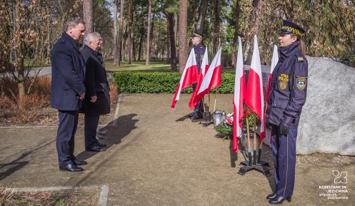 Trzech mężczyzn ubranych na czarno stoi przed pomnikiem katyńskim – dużym kamieniem, na którym wisi granitowa tablica. Pod tablicą leżą kwiaty i palą się znicze. 