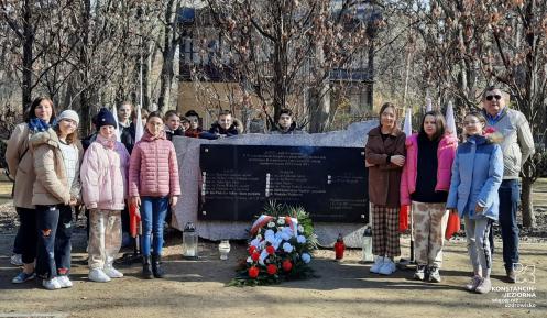 Grupka dzieci oraz dorosłych stoi obok pomnika katyńskiego – dużego kamienia, na którym wisi granitowa tablica. Pod tablicą leżą kwiaty i palą się znicze.
