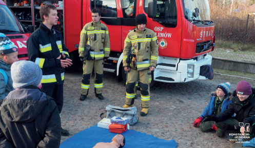 Trzech stojących strażaków na tle wozu strażackiego. Dookoła grupa młodzieży, na ziemi leży niebieska mata, a na niej manekin do ćwiczeń z pierwszej pomocy.