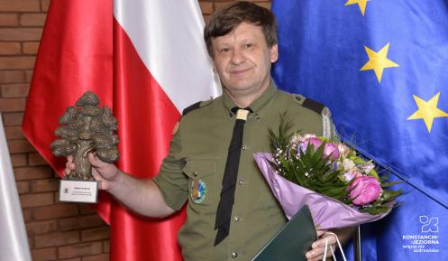 Uśmiechnięty mężczyzna w mundurze harcerskim w jednej ręce trzyma statuetkę w kształcie dębu, a w drugiej – kwiaty, teczkę z dyplomem i torbę podarunkową.