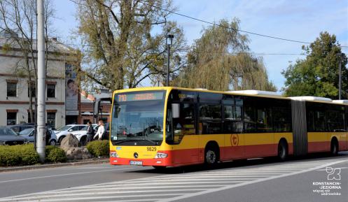 Ulica, po której jedzie czerwono-pomarańczowy autobus komunikacji miejskiej. Nad jego przednią szybą wyświetla się napis: 710 Konstancin-Jeziorna.