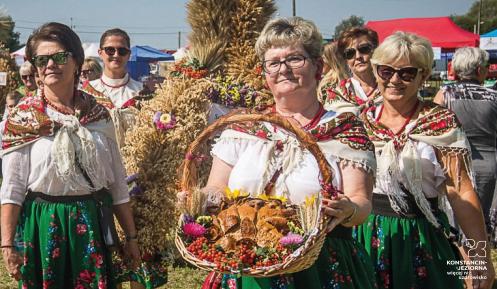 6 kobiet w strojach ludowych. W rękach niosą kosze wiklinowe wypełnione kwiatami i chlebem. 