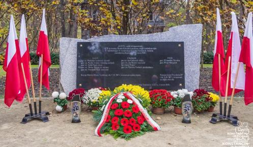 Pośrodku czarna tablica granitowa z tekstem umocowana na prostokątnym kamieniu, po lewej i prawej stronie stoją flagi narodowe, przed tablicą leżą kwiaty i wiązanki oraz palą się znicze