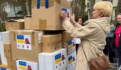 Kobieta w krótkich włosach nakleja kartkę na jeden ze stojących przed nią kartonów z darami medycznymi.