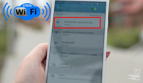 Ręka trzymająca smartfona, na ekranie lista dostępnych sieci wifi, czerwoną ramką zaznaczona sieć o nazwie Konstancin-Jeziorna Free, w górnym lewym roku napis strefa wifi.