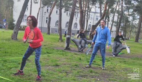 Dwie osoby z rakietkami grają na trawie w crossmintona, w tle dzieci obserwują grę.