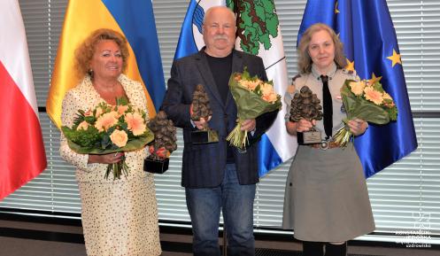 Trzy osoby – dwie kobiety i mężczyzna – stoją na tle flag Polski, Ukrainy, Konstancina-Jeziorny i Unii Europejskiej. W rękach trzymają statuetki dębu oraz kwiaty.