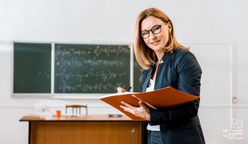  Kobieta – nauczycielka stoi w klasie. W rękach trzyma duży zeszyt. W tle widać tablicę oraz biurko.