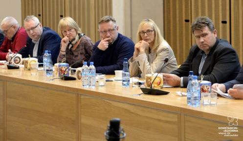 Grupa osób, kobiet i mężczyzn, siedzi przy stole konferencyjnym, na którym leżą dokumenty i stoją mikrofony.