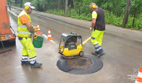 Dwóch robotników na ulicy obok przyczepa. Jeden z robotników prowadzi maszynę dookoła włazu studzienki ulicznej.  