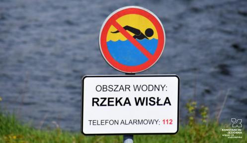 Znak zakazujący kąpieli z tabliczką informacyjną, na której jest tekst: obszar wodny – rzeka Wisła, telefon alarmowy 112. W tle nieostre fale płynącej rzeki.  