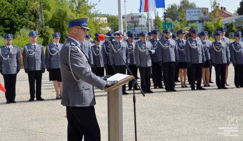 Funkcjonariusze policji w umundurowaniu stoją na baczność w rzędzie. Przed nimi mężczyzna w umundurowaniu przemawia do mikrofonu.